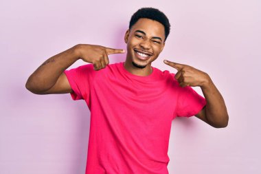 Casua tişörtü giyen, gülümseyen, parmaklarıyla ve ağzıyla işaret eden Afro-Amerikan genç adam. diş sağlığı kavramı. 