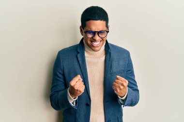 Zarif kıyafetler ve gözlük takan yakışıklı İspanyol bir adam çok mutlu ve heyecanlı bir şekilde kolları havada, gülümseyerek ve başarı için çığlık atarak kazanan bir jest yapıyor. kutlama konsepti. 