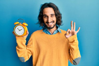 Çalar saati tutan genç İspanyol adam parmaklarıyla imza atıyor, gülümseyerek mükemmel bir sembol oluşturuyor. 