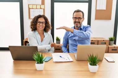 Orta yaşlı İspanyol kadın ve erkek ofiste dizüstü bilgisayarlarıyla oturmuş büyük ve büyük boy işaretleri göstererek el kol hareketi yapıyorlar. Kameraya bakıp gülümsüyor. ölçüm kavramı. 