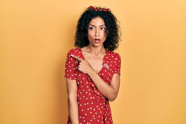 Yazlık elbise giyen genç Latin kız parmaklarını yana doğru göstererek şaşırdı, ağzı açık hayrete düştü.. 