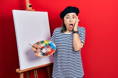 Orta yaşlı İspanyol kadın, resim sehpası üzerine palet çizerek ayakta duruyor. Korkuyor ve şokta. Yüzünde elleri olan, şaşkınlık ve şaşkınlık ifadesi var. 