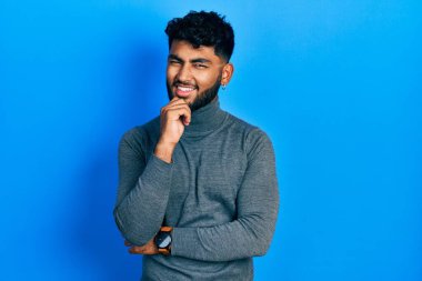 Balıkçı yaka kazak giyen sakallı Arap adam kameraya kendinden emin bakıyor kollarını kavuşturmuş ve çenesini kaldırmış gülümsüyor. Olumlu düşünmek. 