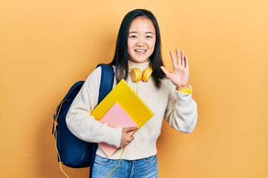Öğrenci çantası ve kitapları tutan genç Çinli kız merhaba diyor mutlu ve gülümsüyor, dostça bir hoş geldin jesti. 