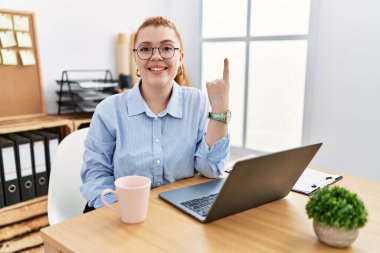 Ofiste çalışan genç kızıl saçlı kadın bilgisayarını kullanarak bir numaralı parmağını gösterip gülümserken kendinden emin ve mutlu.. 