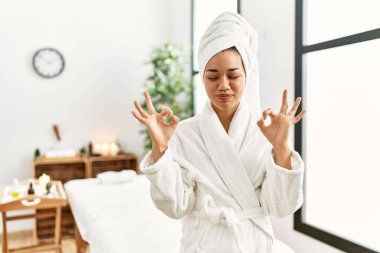 Havlu ve bornoz giymiş genç esmer kadın güzellik merkezinde dikiliyor rahatlıyor ve gözleri kapalı gülümsüyor parmaklarıyla meditasyon hareketi yapıyor. yoga konsepti. 