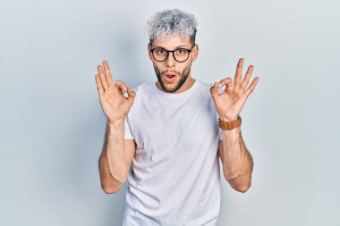Modern boyalı saçlı, beyaz tişörtlü ve gözlüklü, şaşırmış ve şok olmuş genç İspanyol adam parmaklarıyla onay sembolü yapıyor. çılgın ifade 