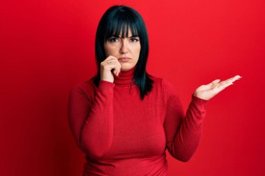 Avuç içleri açık, elinde şüpheci ve gergin bir şey tutan, sorun yüzünden kaşlarını çatan İspanyol bir kadın. negatif kişi. 