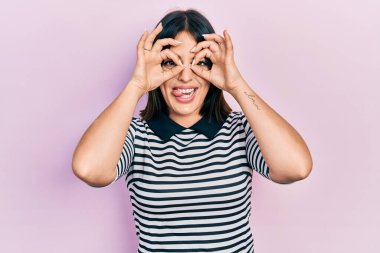 Günlük kıyafetler giyen genç İspanyol kadın dürbün gibi jest yapıyor, gözleri parmaklarından bakıyor. çılgın ifade. 