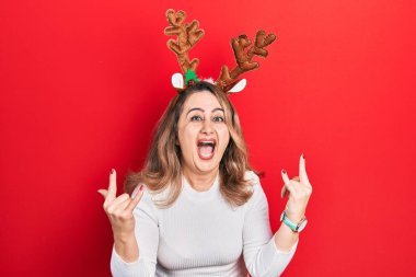 Orta yaşlı beyaz kadın şirin Noel geyiği boynuzları takıyor çılgın bir ifadeyle bağırıyor elleri havada rock sembolü yapıyor. Müzik yıldızı. ağır kavram. 