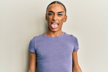 Makyaj yapan, uzun saçlı, sıradan tişörtlü, dilini komik bir ifadeyle dışarı çıkaran İspanyol bir adam. duygu kavramı. 