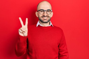 Genç kel bir adam günlük kıyafetler ve gözlük takıyor. İki numaralı parmaklarıyla işaret ediyor. Kendinden emin ve mutlu bir şekilde gülümsüyor.. 