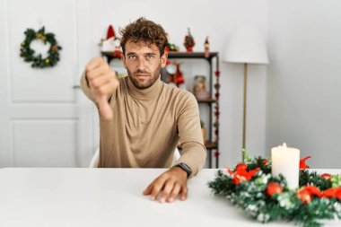 Yılbaşı süslemesi sırasında masanın üzerinde oturan sakallı yakışıklı genç adam mutsuz ve kızgın görünüyor. Reddedilmiş ve olumsuz jestler yapıyor. kötü ifade. 