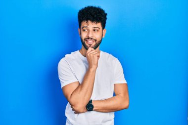 Sakallı, beyaz tişörtlü genç bir adam kameraya kendinden emin bir şekilde bakıyor kollarını kavuşturmuş ve çenesini kaldırmış gülümsüyor. Olumlu düşünmek. 