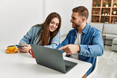 Genç beyaz çift kahvaltılarını yapıyorlar ve evde dizüstü bilgisayar ve akıllı telefon kullanıyorlar..