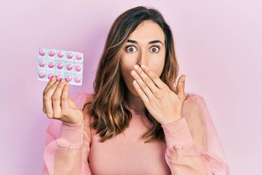 Genç İspanyol kız elinde haplarla ağzını eliyle kapatıyor, şok geçiriyor ve hata yapmaktan korkuyor. şaşırmış ifade 