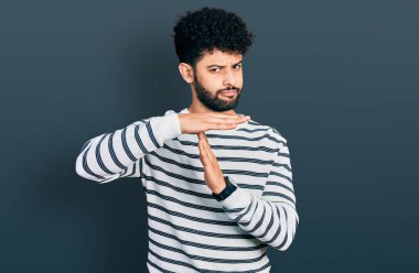 Sakallı, sıradan çizgili süveter giyen, el kol hareketi yapan, hayal kırıklığına uğramış ve ciddi yüzlü genç bir Arap. 