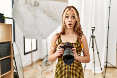 Fotoğraf stüdyosunda elinde profesyonel kamerayla gezen beyaz bir genç kız ağzı açık kalmış, korkmuş ve şaşkın bir şekilde şaşırmış. 