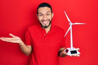 Sakallı İspanyol bir adam yenilenebilir elektrik için güneş değirmeni tutuyor başarıyı mutlu bir gülümsemeyle kutluyor ve kazanan el kaldırarak ifade ediyor 
