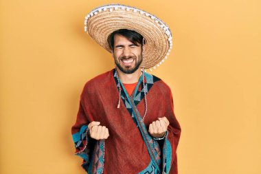 Meksikalı şapkalı genç İspanyol adam çok mutlu ve heyecanlı. Kolları havada, gülümseyen ve başarı için bağıran kazanan bir jest yapıyor. kutlama konsepti. 
