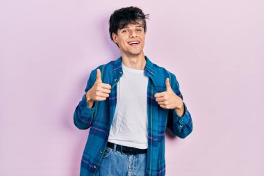 Yakışıklı hippi genç adam, günlük beyaz tişört ve klasik tişört giyiyor. Başarı işareti el hareketleriyle olumlu jest yapıyor. Gülümsüyor ve mutlu. Neşeli ifade ve kazanan jest. 