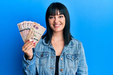 Genç İspanyol kadın Suudi Arabistan riyal paralarını tutuyor. Pozitif ve mutlu görünüyor. Kendine güvenen bir gülümsemeyle dişlerini gösteriyor. 