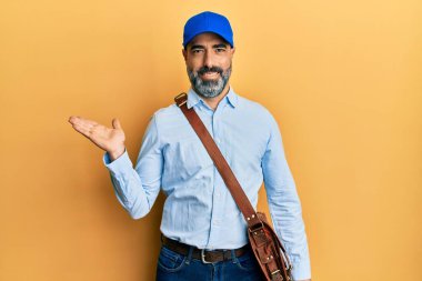 Orta yaşlı, sakallı, gri saçlı kurye şapkası takan neşeli bir şekilde gülümseyen ve avuç içi kameraya bakan bir adam.. 