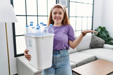 Geri dönüşüm sepetini elinde tutan, elinde plastik şişelerle neşeli bir şekilde gülümseyen ve avuç içi kameraya bakan genç kızıl saçlı kadın.. 