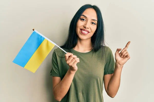 Mladá Hispánka Nosí Ukrajinskou Vlajku Šťastně Ukazuje Rukou Prstem Stranu — Stock fotografie