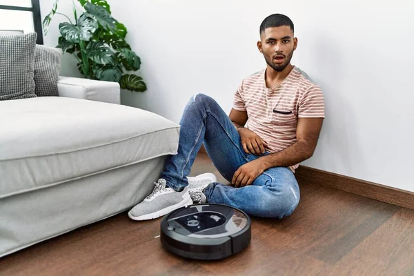 Young indian man sitting at home by vacuum robot scared and amazed with open mouth for surprise, disbelief face