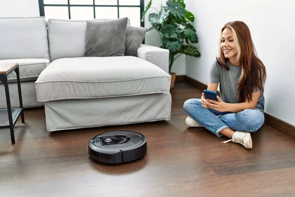 Evde yerde oturan akıllı telefonuyla robot elektrik süpürgesini kontrol eden beyaz bir kız..