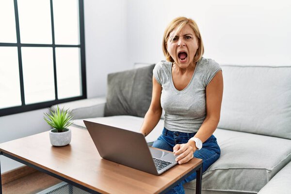 Блондинка средних лет использует ноутбук дома злой и безумный крик разочарованный и разъяренный, кричащий от гнева. гнев и агрессивная концепция. 