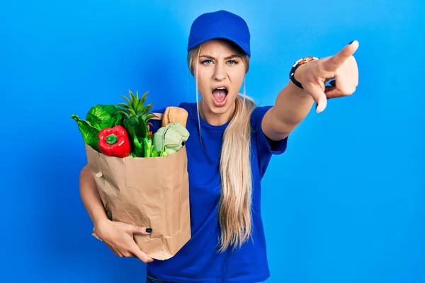 若いですCaucasian女性身に着けています宅配便制服で食料品からスーパーマーケットを指して不満とカメラにフラストレーション 怒りと激怒であなた — ストック写真