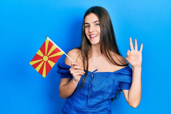 Молодая брюнетка подросток держит македонский флаг делает хорошо знак с пальцами, улыбаясь дружелюбный жест отличный символ 