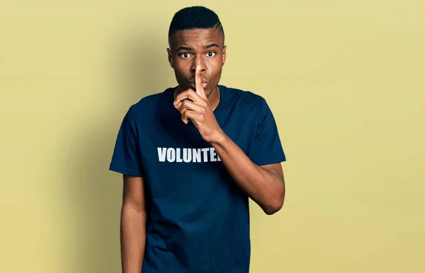 ボランティアのTシャツを着た若いアフリカ系アメリカ人男性が唇に指で静かになるように求めています 沈黙と秘密の概念 — ストック写真