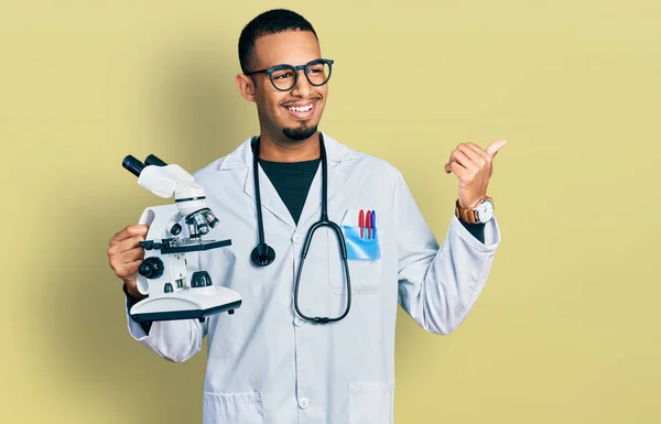 Jovem Afro Americano Vestindo Uniforme Cientista Segurando Microscópio Apontando Polegar — Fotografia de Stock