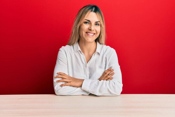 Молодая белая женщина в повседневной одежде сидит на столе счастливое лицо, улыбаясь скрещенными руками, глядя в камеру. позитивный человек. 