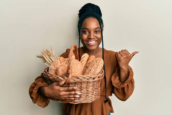African american woman with braided hair holding wicker basket with bread pointing thumb up to the side smiling happy with open mouth
