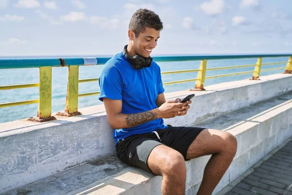 Young latin man smiling happy using headphones and smartphone at the beach.