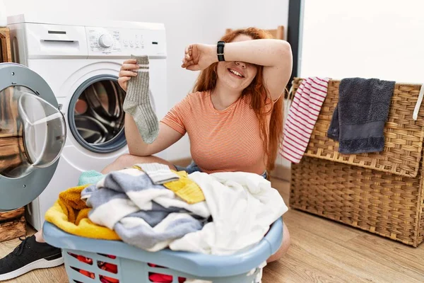 若い赤毛の女性が洗濯機に汚れた洗濯機を入れて陽気に顔を示す手で竹を覗かせて笑っている びっくりして興奮しました — ストック写真