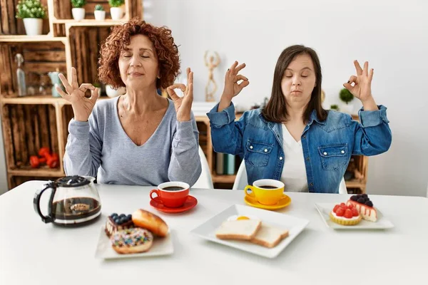 母親とダウン症候群の娘の家庭で朝食を食べに座って家族がリラックスし 目を閉じて指で瞑想ジェスチャーを行う笑顔 ヨガのコンセプト — ストック写真