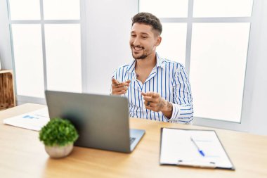 Ofiste çalışan sakallı genç yakışıklı adam bilgisayarla kameraya mutlu ve komik suratını gösteriyor. iyi enerji ve titreşimler. 