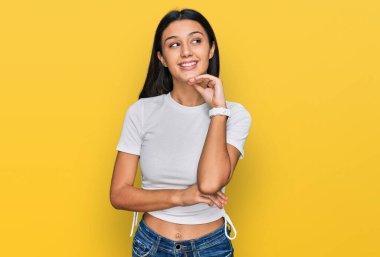 Elleri çenesinde, gündelik beyaz tişört giyen, soru soran genç İspanyol kız, düşünceli yüz ifadesi. Gülümseyen ve düşünceli bir yüz. şüphe kavramı. 