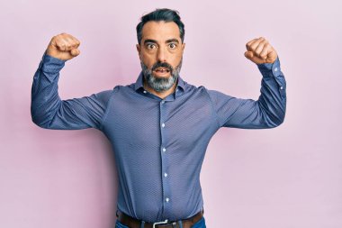 Orta yaşlı, sakallı, gri saçlı, iş kıyafetleri giyen, gururla gülümseyen kol kasları gösteren bir adam. fitness konsepti. 