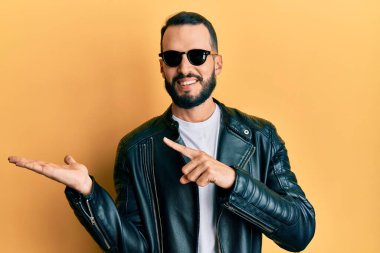 Siyah deri ceket ve güneş gözlüğü takan sakallı genç adam kameraya hayret ediyor ve gülümsüyor.. 