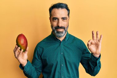 Orta yaşlı, sakallı ve gri saçlı bir adam mango tutuyor. Parmaklarıyla imza atıyor, gülümsüyor. Mükemmel bir sembol. 