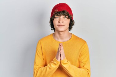 Yakışıklı genç adam yün şapka takıyor ellerle dua ediyor af diliyor kendinden emin bir şekilde gülümsüyor.. 