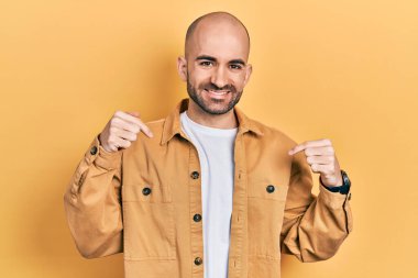 Gündelik kıyafetler giyen genç kel bir adam. Kendine güvenen, yüzünde gülümseme olan, kendini gururlu ve mutlu gösteren biri.. 