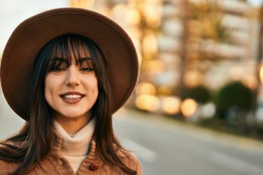 Kışlık şapkalı esmer kadın gün batımında şehirde gülümsüyor.