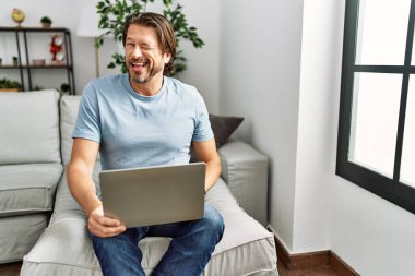 Kanepede bilgisayar kullanan yakışıklı orta yaşlı bir adam kameraya seksi bir ifadeyle bakıyor, neşeli ve mutlu bir yüz.. 